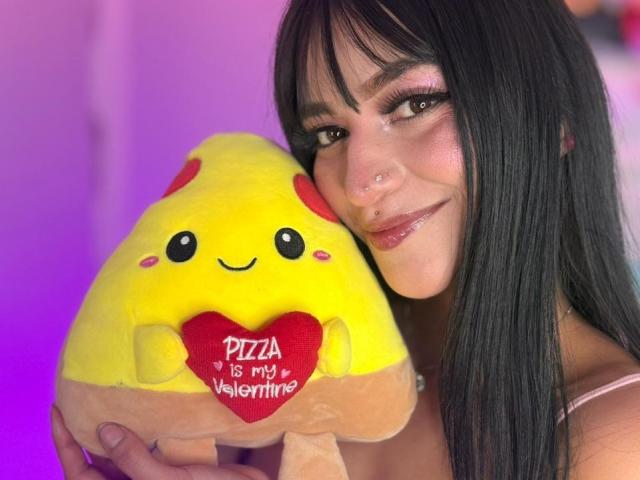 LindaWatsong with black hair, nose ring, and pink lipstick, holding a yellow pizza stuffed toy with red heart and "Pizza is my Valentine" text, smiling.
