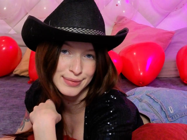 MeganSolar, avec ses cheveux bruns, portant un chapeau de cowboy noir et un haut noir pailleté, souriante, avec un visage constellé de taches de rousseur, des ballons rouges, un jean bleu et un coussin rose en arrière-plan..