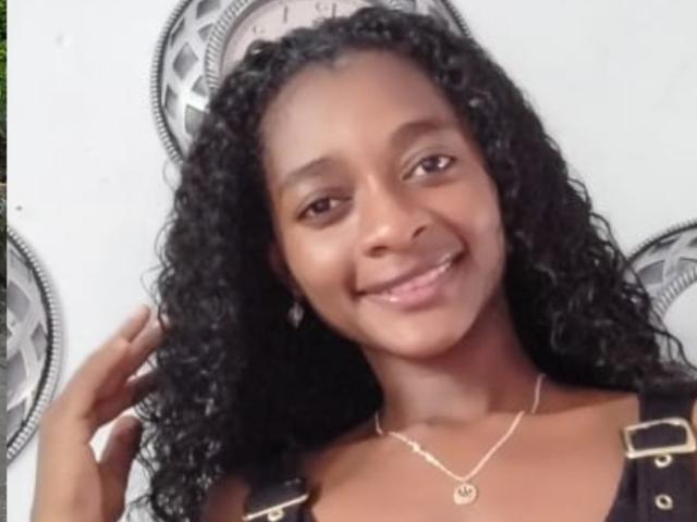 Natix has dark curly hair, brown eyes, and a smile showing white teeth. She wears a gold necklace and black straps with gold buckles on her shoulders.