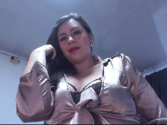 DakotaGray with dark hair, red lipstick, wearing a shiny pink satin top tied at the front, black lace bra visible, raising her right arm, smiling, in a low-angle shot.