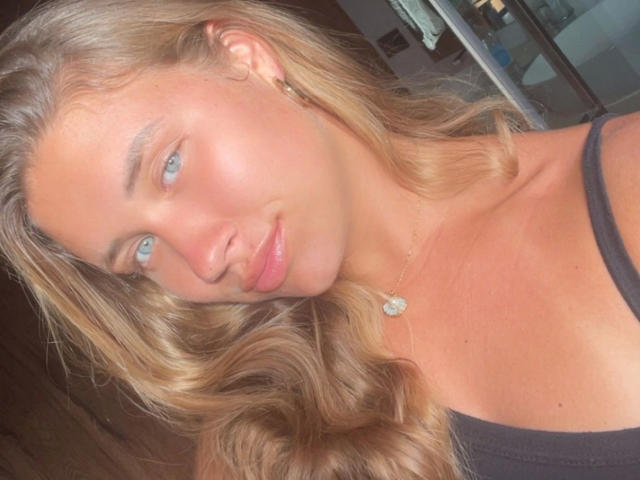 Juicybooty6-hot has light blue eyes and wavy blonde hair that falls to her shoulders. She's wearing a black tank top and a gold necklace with a small, round pendant. Her skin is smooth and slightly shiny, with a subtle pinkish hue. She's looking to the left with a slight, relaxed smile. The lighting is soft and warm, highlighting her facial features. Her ear is adorned with a small gold hoop earring. The image is close-up, focusing on her face and upper chest.