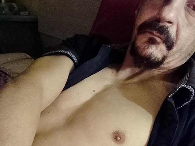 Dragone69 with a dark mustache, open black shirt, and exposed chest, showing small, perky tits and a light skin tone, lying down in dim lighting.