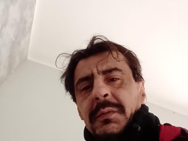 Dragone69 has a messy black mustache, slightly wavy dark hair, and a serious expression, wearing a black and red top, in a low-angle selfie.