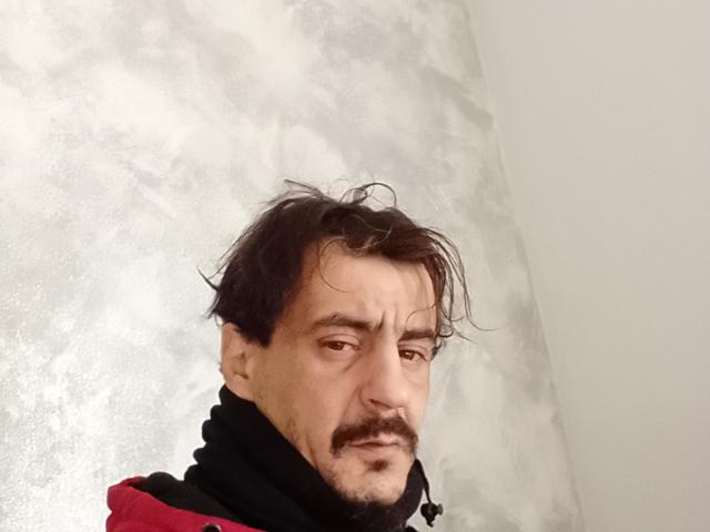 Dragone69 has messy black hair, a mustache, and a slightly worried expression. He's wearing a black and red hoodie with a black scarf. His nose is small.