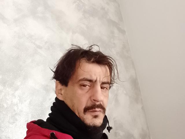 Dragone69 with messy brown hair, mustache, and slight smirk, wearing a black and red jacket, looking tired. Grey, textured wall in the background.