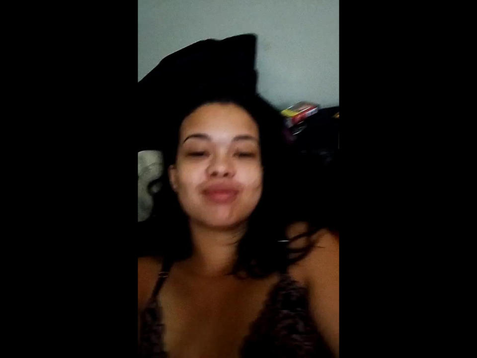 MariJapinha with dark hair, smiling, wearing a lace top, has small tits, light brown skin, and is lying down with black hair covering part of her face.
