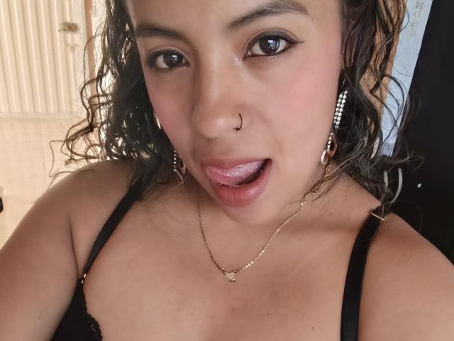 MeganFerrerr has curly brown hair, a nose ring, and is sticking out her tongue with her lips slightly parted. She's wearing black lace lingerie and a gold necklace.