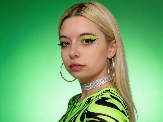 Reginia with blonde hair, green eye makeup, silver glitter on cheek, hoop earrings, chandelier necklace, shiny pink lips, green and black zebra-patterned top, looking to the side.