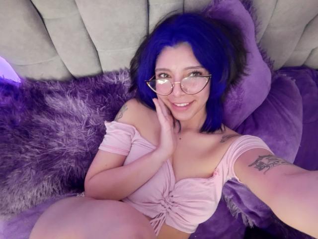 DarckVelvet with blue hair, glasses, smiling, pink off-shoulder top, hand on cheek, visible tattoo on right arm, lying on purple furry and velvet pillows, cute and sexy.