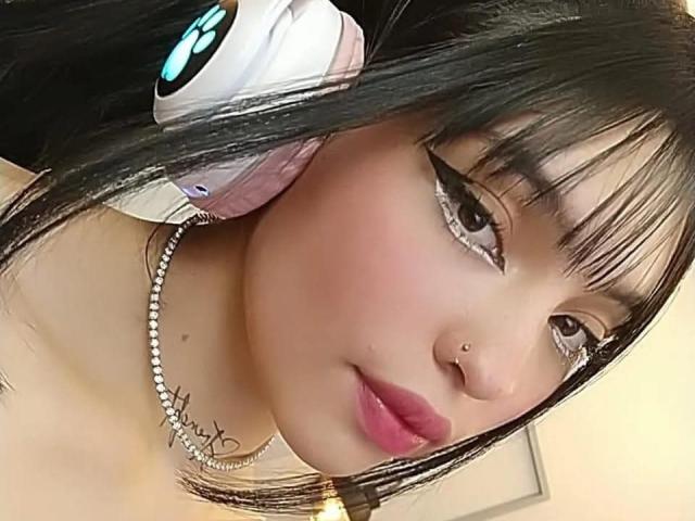 KuroBabee with black hair, wearing pink headphones, has a nose piercing and thick eyeliner, wearing a pearl necklace, and pink lipstick, showing off her cute face and tits.