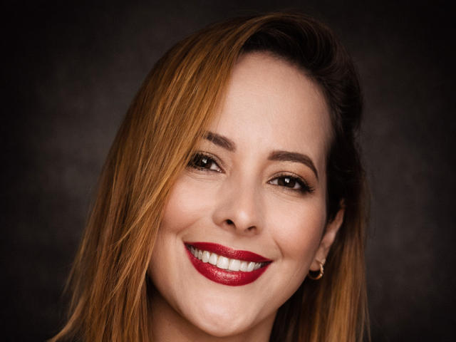 Close-up of SaritaVelez with light brown, shoulder-length hair, slightly parted to the left. She has fair skin, dark brown eyebrows, and brown eyes. She's smiling with red lipstick, showing white, even teeth. She wears small gold earrings. The image has a dark, blurred background. Her hair texture is smooth, and the lighting highlights her facial features. The overall style is realistic and focused on her facial expression. The photograph uses sharp detail and subtle shadows to emphasize her smile.