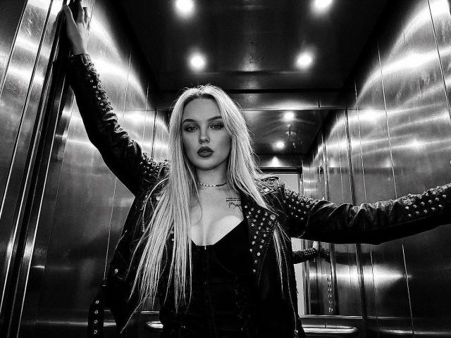 JaneOSweetAh with long blonde hair, dark lipstick, and studded black leather jacket, arms spread, standing in a metallic elevator, looking straight at camera, monochrome, serious expression.
