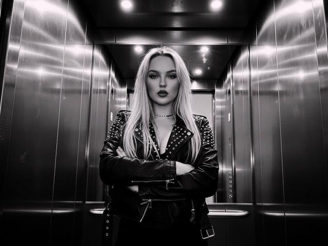 JaneOSweetAh stands in a metal elevator, arms crossed, wearing a black leather jacket with studs, choker, and long blonde hair, looking intense with dark lipstick. Black-and-white photo.
