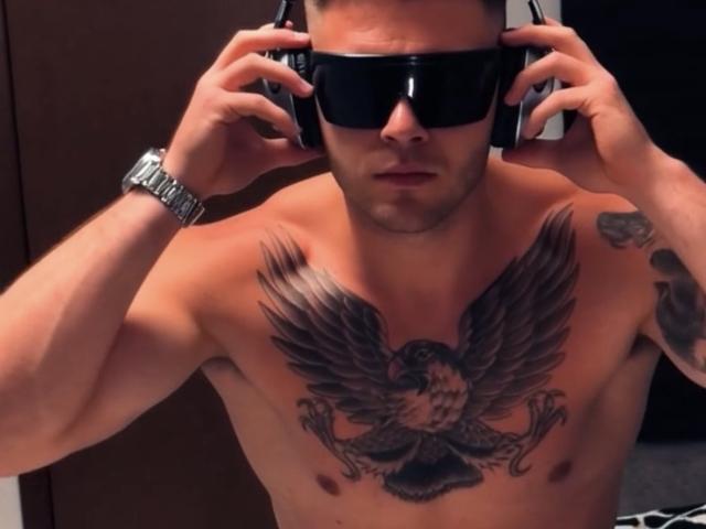 Shirtless Antzo with a muscular build, wearing black sunglasses, and large headphones. He has a detailed black eagle tattoo with outstretched wings covering his chest. Antzo's right arm has a tattoo on the outer side. He wears a silver watch on his left wrist. The image has a slight shadow, emphasizing his upper body. The lighting highlights his tattoos and muscular definition. His expression is focused. The overall style is modern with a slight gritty, edgy vibe.