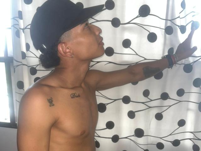 ChrisSexi, shirtless with a black cap, tattoo on right shoulder, arm raised, touching curtain with black circles. He's wearing a bracelet and has short, curly hair.
