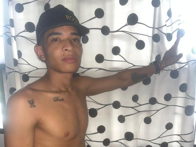 ChrisSexi, shirtless, wearing black cap, tattoos on left shoulder, right arm tattoos, brown skin, standing against polka dot curtain, right arm raised, looking at camera, pouty expression.