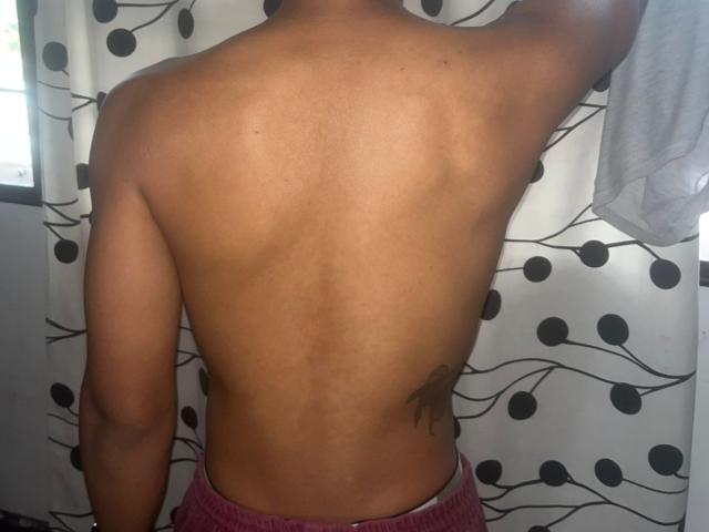 ChrisSexi stands with his back to the camera, showing a toned, muscular back with a visible spine and small tattoo on the right side. He has medium brown skin and is shirtless, wearing pink pants. His right arm is raised, holding a white shirt. The background has a white curtain with black circular patterns. The lighting is natural. The image is clear, showing the texture of ChrisSexi's skin and muscle definition.