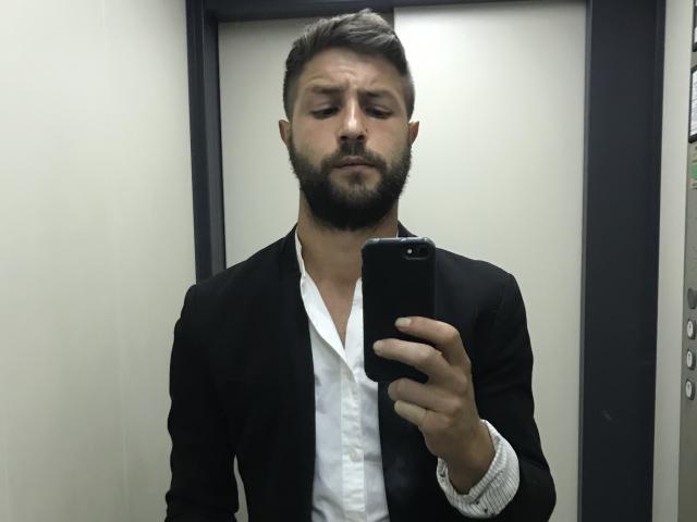 Antzo with a beard, brown hair, and a black suit, holding a phone, looking confused, in a mirror selfie. His white shirt is partially visible.