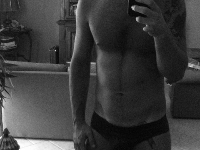 Black-and-white selfie of Antzo, a shirtless, muscular man with a defined chest and abs, wearing dark underwear. Antzo holds a phone in his right hand, partially covering his face. His left hand is relaxed at his side. The image has a grainy texture, and the background shows a couch, a plant, and a bookshelf. Antzo's skin is light, and his hair is dark and wavy. The focus is on Antzo's torso.