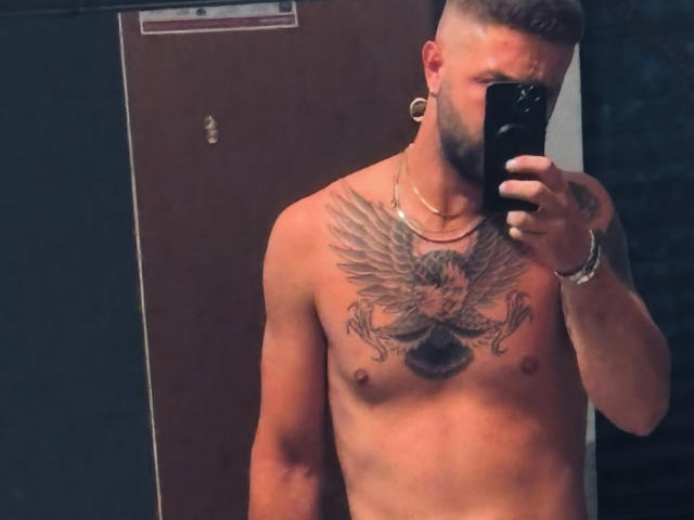 Antzo with a shaved head and beard, holding a phone, shirtless, showing a large eagle tattoo on his chest, wearing gold hoop earrings and a bracelet.