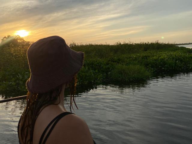 KataSofia with wet brown hair, wearing a black tank top and brown bucket hat, stands in a river at sunset, facing the water with sun on her back.