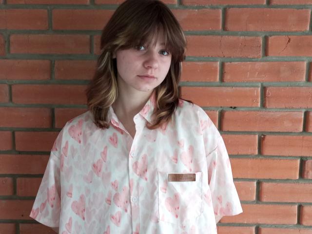 AnnaWestt with shoulder-length brown hair, wearing a pink heart-patterned button-up shirt, standing against a red brick wall, looking slightly bored. Shit, she looks like a nerd.