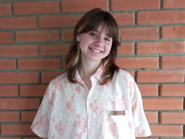 AnnaWestt with brown hair, smiling, wearing a pink floral button-up shirt, standing against a red brick wall. She has a small name tag on the shirt's chest.