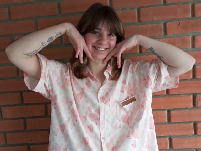 AnnaWestt with brown hair, smiling, wearing a pink floral shirt, hands on cheeks, showing tattoos on both arms, standing against a red brick wall.