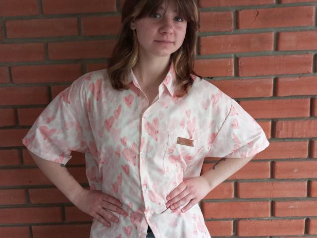 AnnaWestt stands with hands on hips, wearing a pink and white butterfly-patterned shirt, brick wall in background, brown hair, slightly smiling, white skin, small brown name tag.