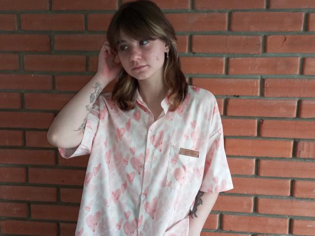 AnnaWestt with light brown hair, wearing a pink heart-patterned shirt, standing against a brick wall, hand in her hair, tattoos on her right arm, looking to the side.