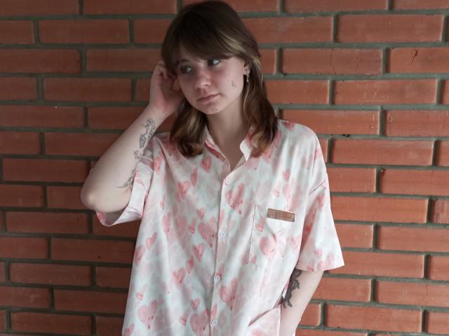 AnnaWestt with brown hair, wearing a pink floral button-up shirt, standing against a brick wall, hand touching her head, showing tattoos on her arm and chest.