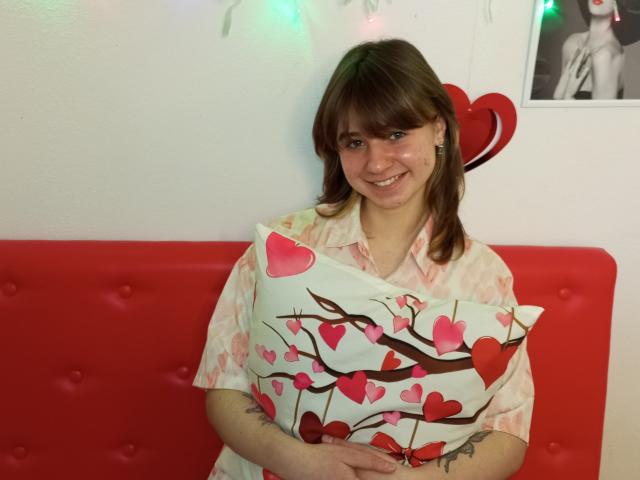 AnnaWestt with brown hair and bangs, wearing a pink checkered shirt, holding a pillow with red and pink heart patterns, smiling, standing in front of a red cushioned wall.