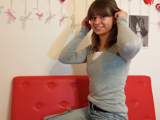 AnnaWestt with brown hair, smiling, wearing a gray long-sleeve top, blue jeans, and black belt, sitting on a red tufted bench, hands in hair.