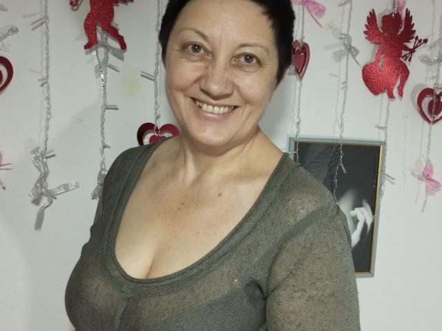 EmilyLlove, middle-aged woman with short black hair, smiling, wearing a sheer green top that reveals her small boobs, standing in front of a wall with red and silver ornaments.