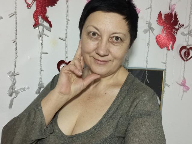 EmilyLlove with short black hair, gray shirt, and cleavage, smiling, holding her cheek, medium tits, fair skin, brown eyes, in a room with red and silver decorations.