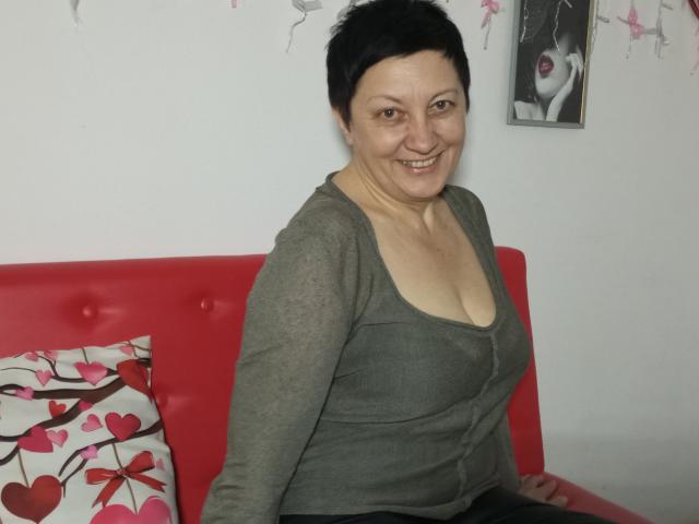 EmilyLlove has short black hair, a smiling face, and small tits in a green, long-sleeve, low-cut top, sitting on a red couch with pink heart pillows.