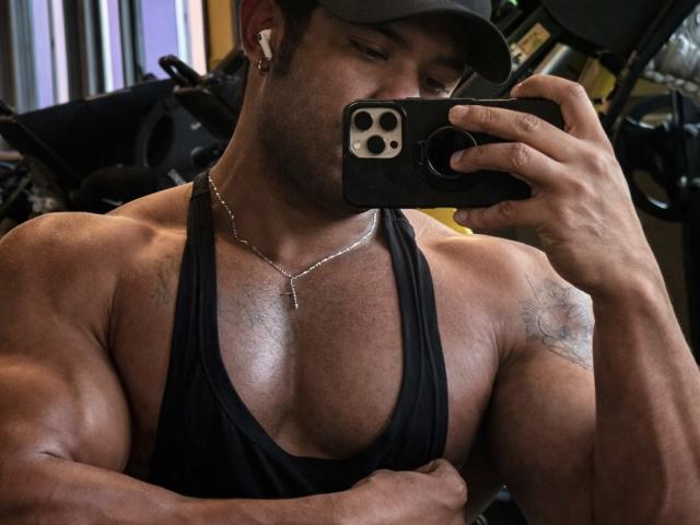 LeonHrz, muscular, tan-skinned, wearing a black tank top and cap, taking a selfie in a gym with his right hand, showing his defined chest and arm tattoos.