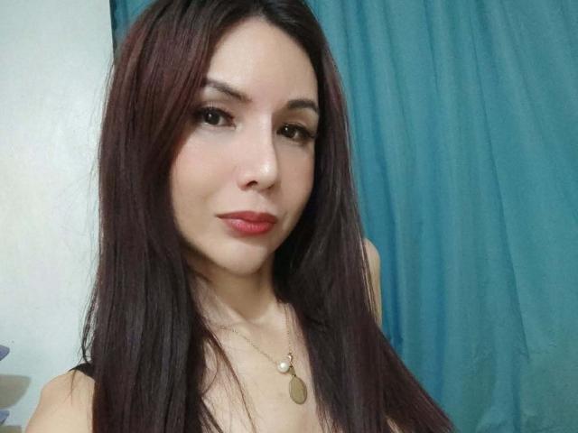 MissHotAyaLove with long dark brown hair, red lipstick, and a small gold necklace, smiling softly at the camera, wearing a black top, showing her tits slightly.