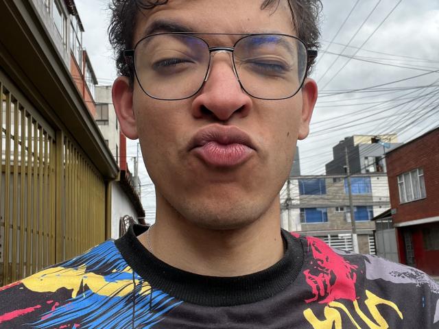 MiloJhones with puffy lips, wearing glasses, and a colorful shirt, has curly hair and a slightly tan complexion, making a silly face in a street setting.