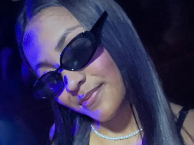NaomiRios with long gray hair, wearing black sunglasses and a pearl necklace, smiling with her head tilted slightly to the side, under blue lighting.