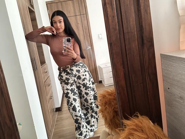 Soranaa with long black hair, brown top, and black/white floral pants, taking a mirror selfie. She's holding a phone with a pink case, showing off her ass.