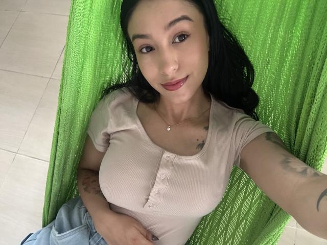 Soranaa with dark hair, tan ribbed shirt, buttoned up, showing big tits, lying on green fabric, smiling, hand on belly, tattoo on arm, wearing light jeans.