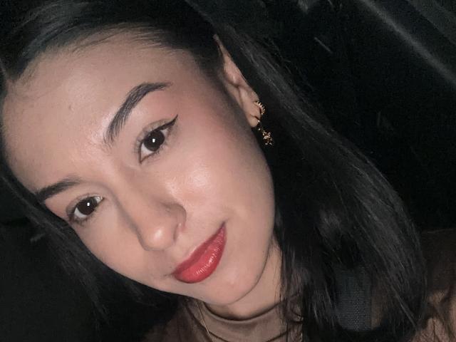 Soranaa with dark hair, red lipstick, and black eyeliner, has a cute smile, wearing gold earrings, in a dark car, with a seatbelt visible.
