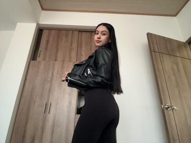 Soranaa a de longs cheveux noirs, elle porte une veste en cuir noir et un pantalon noir taille haute, qui met en valeur ses fesses. Elle se tient dans une pièce avec des portes en bois et un mur blanc..