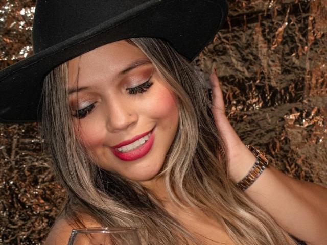 KedallBennet, close-up portrait. Wearing a black hat, she has long, wavy, blonde hair with dark roots, and bright pink lipstick. Her eyes are closed with pink eyeshadow and long, thick eyelashes. She's smiling slightly with her right hand touching her hair. A gold wristwatch is visible on her left wrist. Background is blurred, featuring brown, dried leaves. Image has warm, natural lighting.