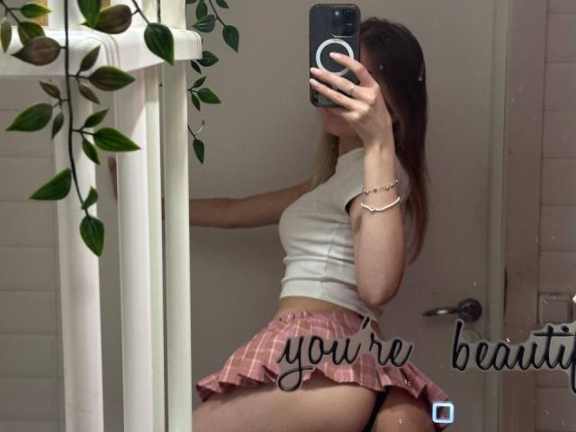 MichaFoxy in a white crop top and pink plaid skirt, showing her ass in a mirror selfie, holding a phone with green leaves in the background.