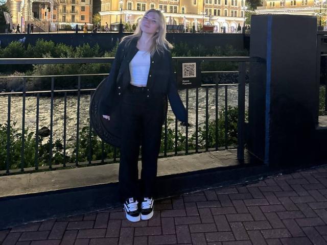 MichaFoxy stands in front of a black metal fence, leaning slightly to the right. She has long blonde hair, wears a black jacket over a white crop top, black high-waisted pants, and black-and-white sneakers. Her right hand holds a black bag. The background shows a river and yellow buildings with trees. The pavement is red brick. The QR code is on the fence. Bright daylight.