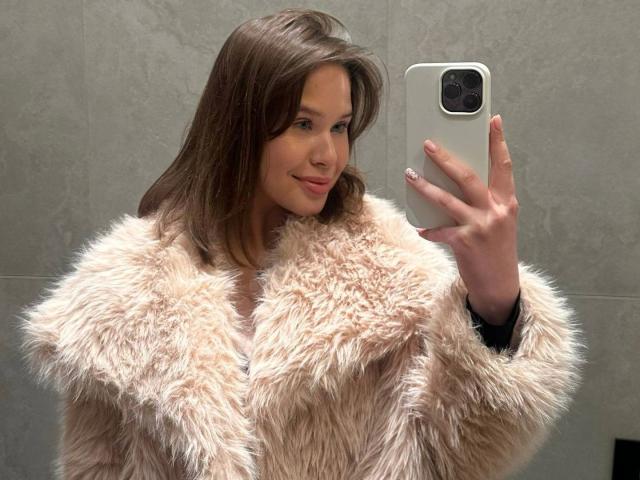 Rosally-hot with brown hair, wearing a fluffy beige fur coat, holding a phone in a mirror selfie, smiling slightly, showing a cute, attractive face, and white nails.