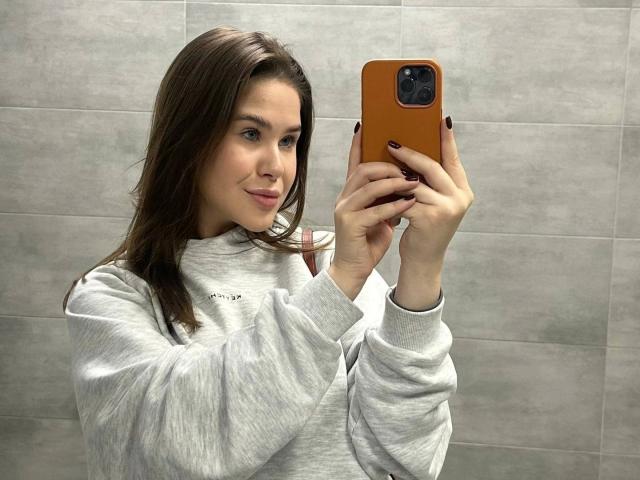 Rosally-hot with brown hair, wearing a gray sweater, taking a selfie with an orange phone, smiling slightly, nails painted black.