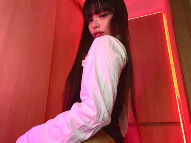 PrincessMiley with long black hair, wearing glasses and a white long-sleeve shirt, sitting with her back to the camera, looking over her shoulder, in red lighting.